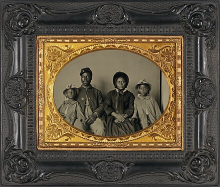 A photograph of an African American Union soldier in uniform posing with his wife and two daughters, circa 1863-1865