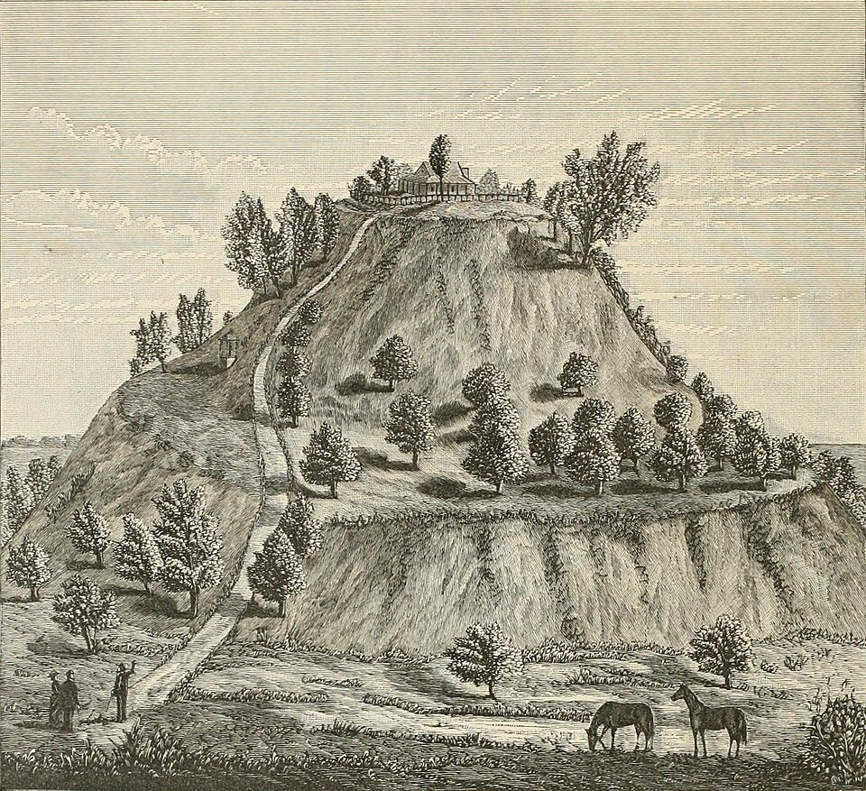 1882 engraving of Monks Mound at Cahokia — a massive terraced earthen mound covered in trees, with a house on its summit, a path winding up the side, and two human figures and grazing horses at the base for scale.