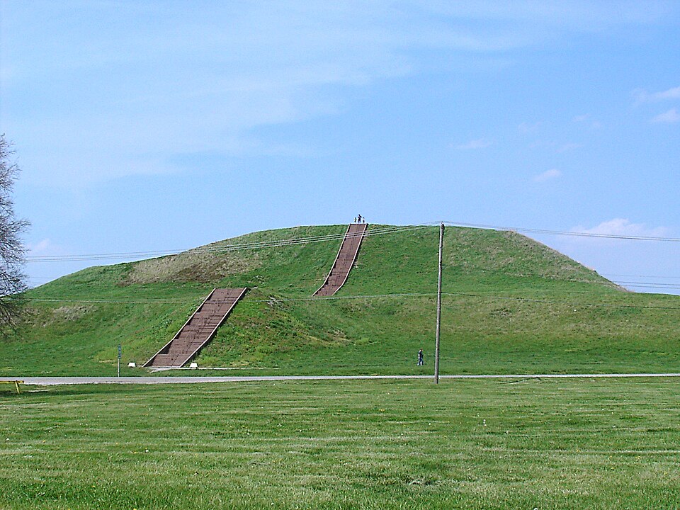 Monks Mound at Cahokia — a massive flat-topped earthen pyramid rising approximately 100 feet, with tiny human figures at its base for scale. The mound covers 14 acres.