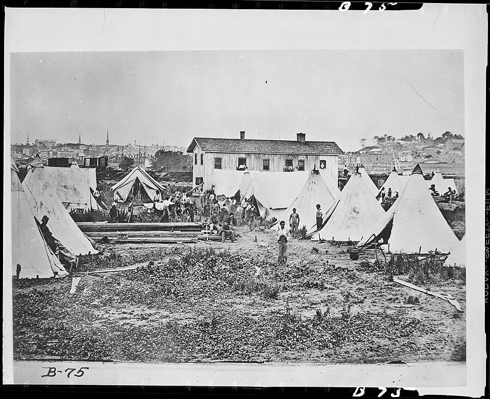 Photograph of formerly enslaved people gathered in a contraband camp near Richmond, Virginia, c. 1865. Rows of simple wooden shelters visible. Men, women, and children looking directly at the camera.